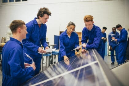 A group of engineering students stand over some solar panels and work on a group project together. They are all wearing blue coveralls.